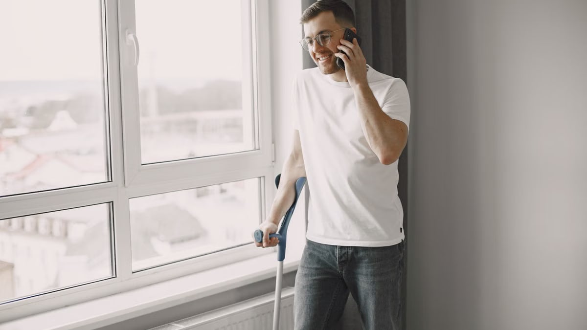 Young man standing by the window on crutches, talking on smartphone with a smile.